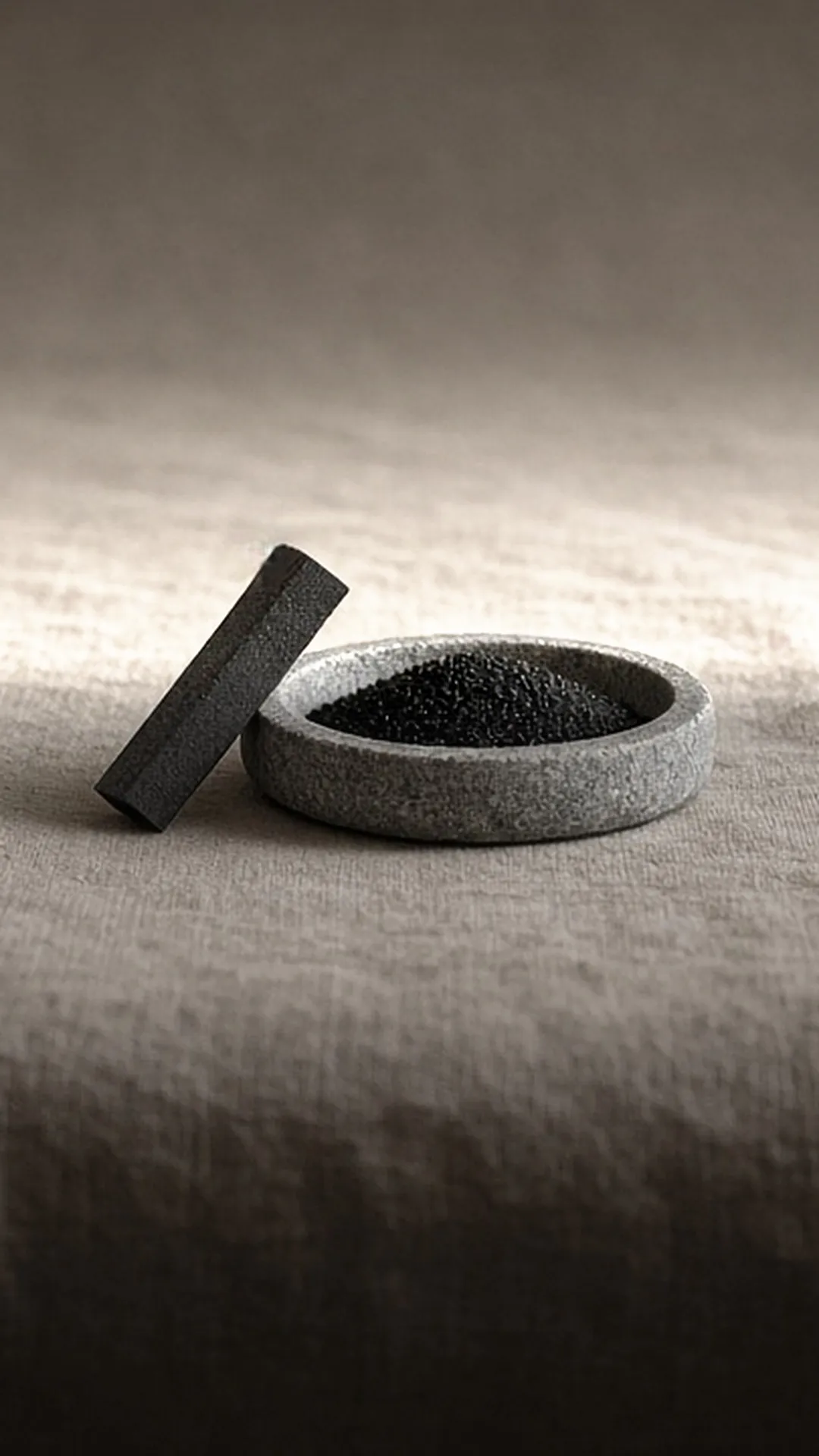 A dark filter block leaning against a shallow stone dish of black granules on grey linen